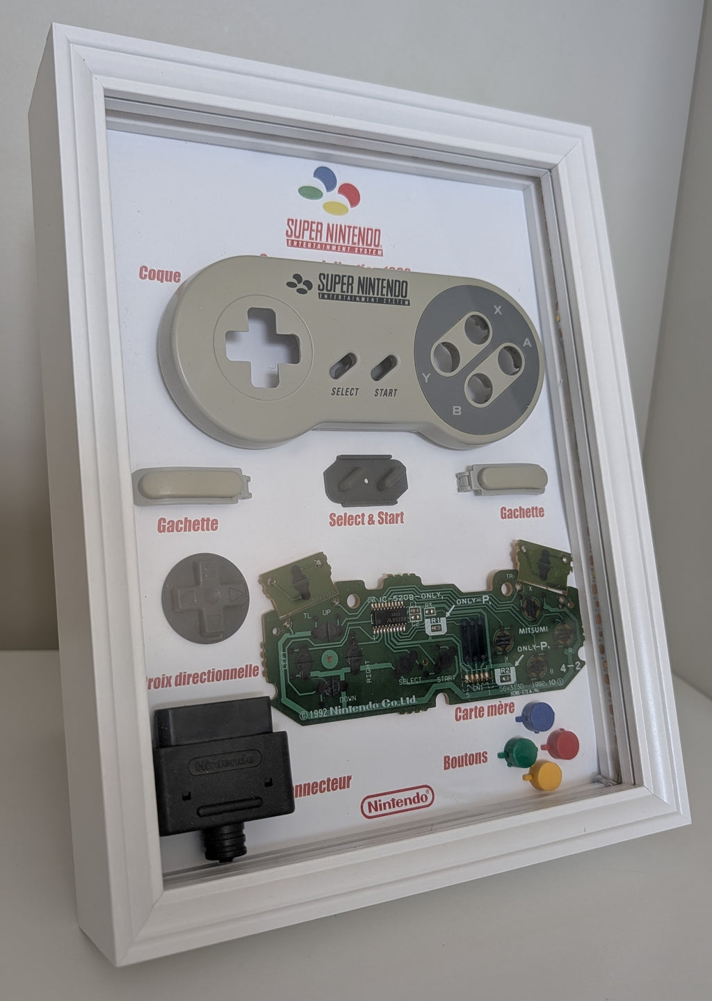 Diorama Super Nintendo avec véritable manette SNES démontée et composants exposés dans un cadre blanc